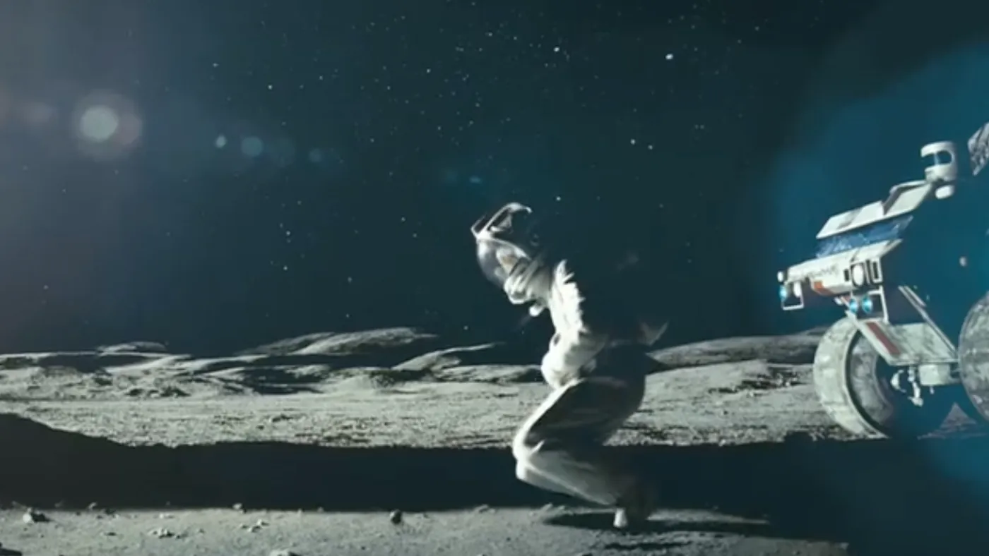 A suited figure kneels on the surface beside a rover
